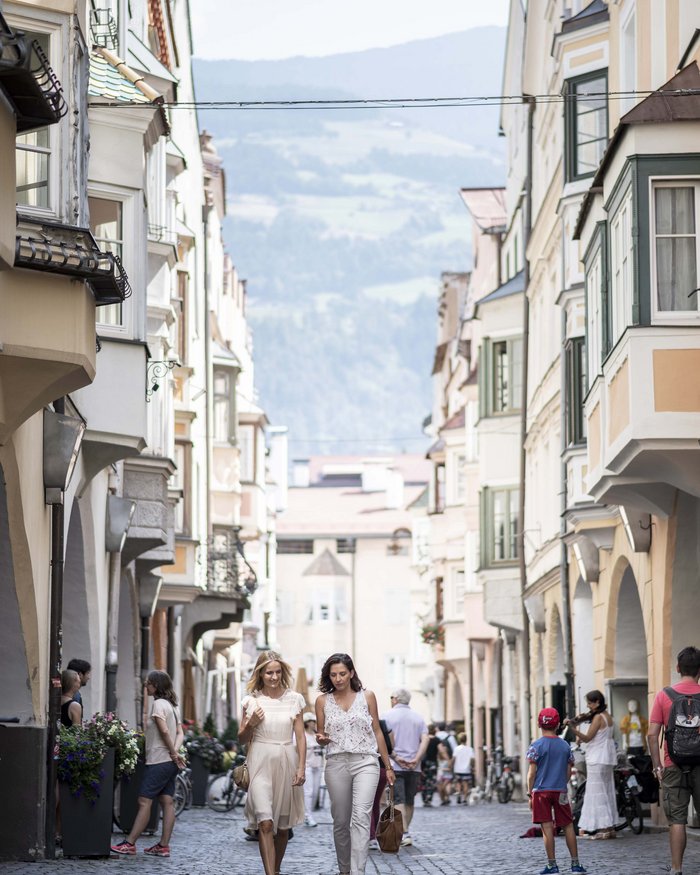 Due donne camminano in una via affollata con edifici storici e montagne sullo sfondo