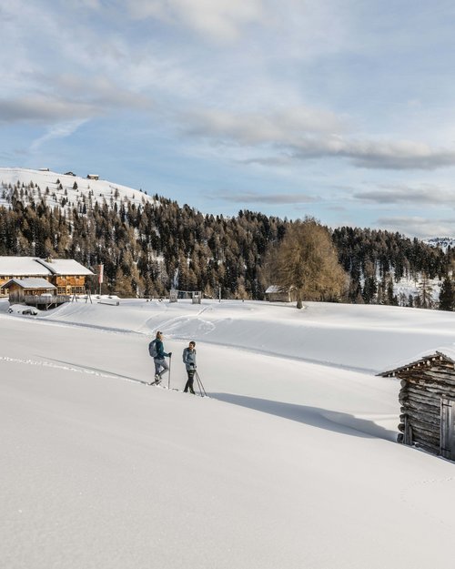 Due persone con racchette da neve in paesaggio montano innevato con baite