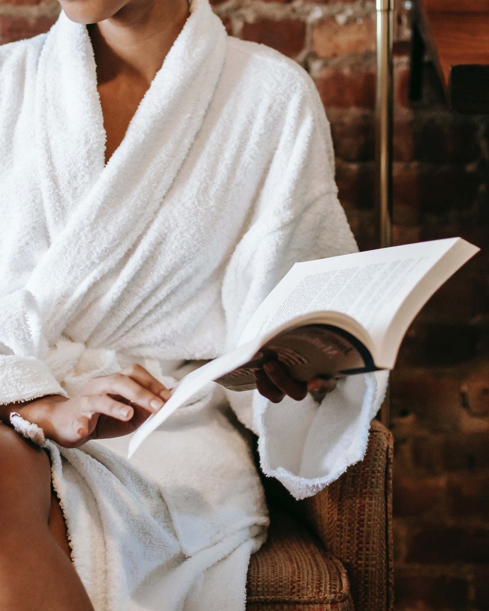 Donna in accappatoio bianco legge un libro seduta davanti a muro di mattoni